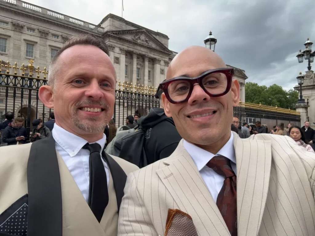 Isaac and his partner stand outside Buckingham Palace, wearing suits.