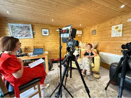 woman in red (Laura Doyle) sitting on a red chair interviewing a Network member a woman in a black top and flower printed pants sitting on a yellow sofa with 2 black cameras on a tripod in the background
